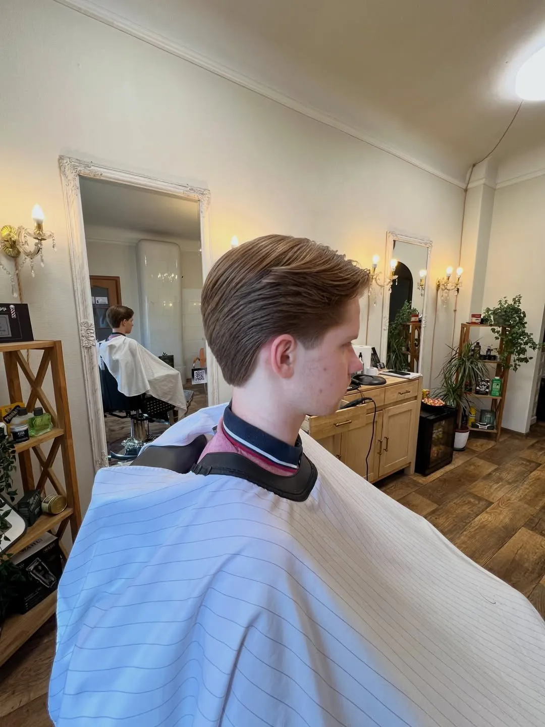 Klients frizieru krēslā ar svaigi ieveidotu, atpakaļ ķemmētu frizūru; fonā gaišs salons ar spoguļiem un sienas lampām